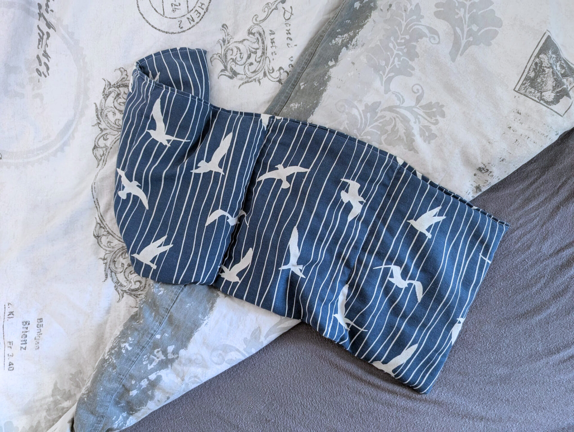 Selbstgemachtes Wärmekissen aus blauem Stoff mit weißen Möwenmustern, in Kammern genäht, liegt auf einem Bett mit gemusterter Bettwäsche.