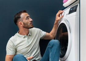 Mann sitzt vor einer Waschmaschine, die stehen geblieben ist, und blickt ratlos auf das Display.