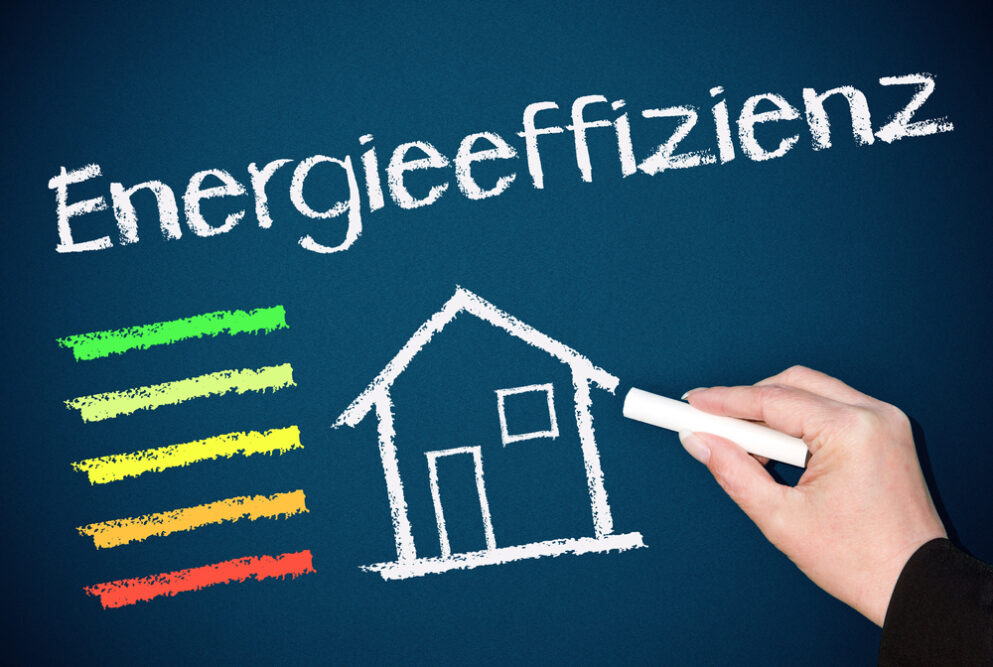 Illustration eines Energieausweises mit farbiger Skala von grün bis rot und einem gezeichneten Haus, das mit Kreide auf eine blaue Tafel gezeichnet wurde. Text 'Energieeffizienz' steht darüber.