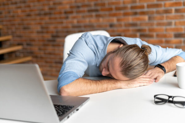 Ein Mann schläft am Schreibtisch mit dem Kopf auf den Händen neben einem Laptop und einer Brille. Doch wie lange soll ein Powernap dauern?