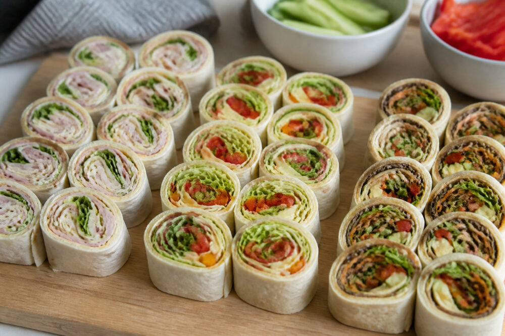 Wrap-Röllchen mit verschiedenen Füllungen auf einem Holzbrett, daneben geschnittenes Gemüse in kleinen Schalen.
