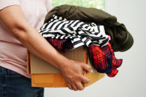 erson trägt einen Karton mit aussortierter Kleidung (Jacke, gestreiftes Shirt, kariertes Hemd) – Symbolbild für Altkleider abgeben und Kleiderspende.