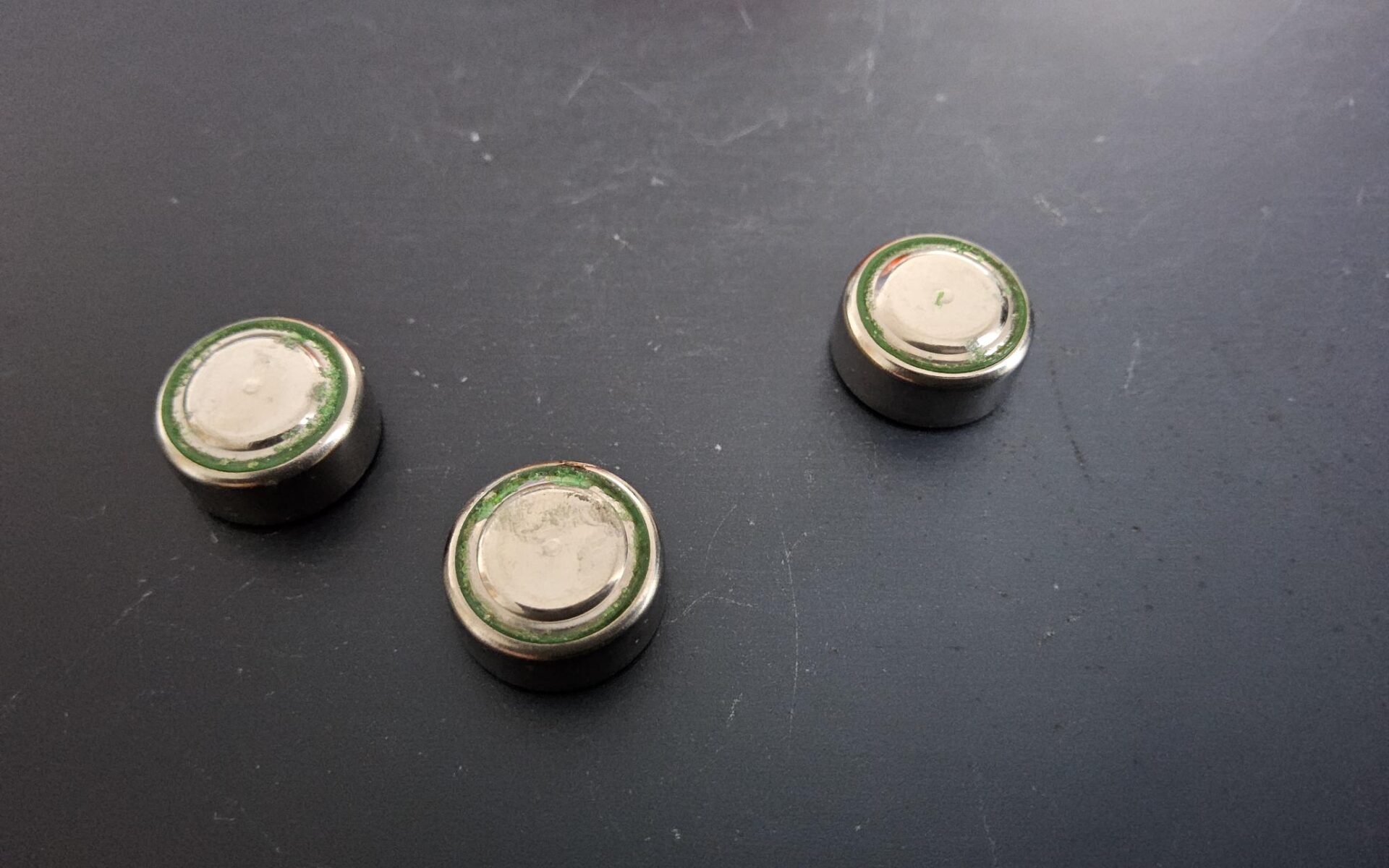 Drei ausgelaufene und korrodierte Knopfzellen (Batterien) liegen mit der Oberseite auf einem dunklen Tisch.