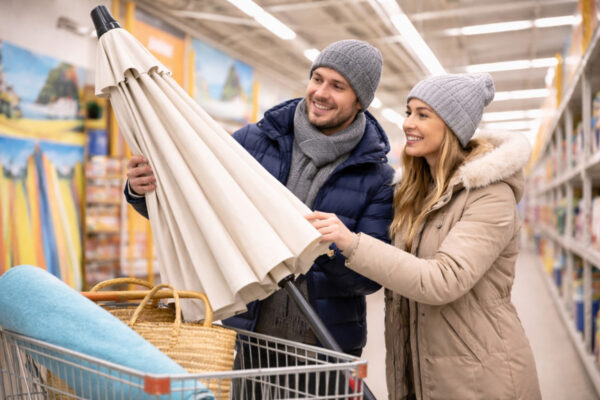 Außerhalb der Saison einkaufen: Paar im Baumarkt in Winterjacken schaut sich einen Sonnenschirm an.