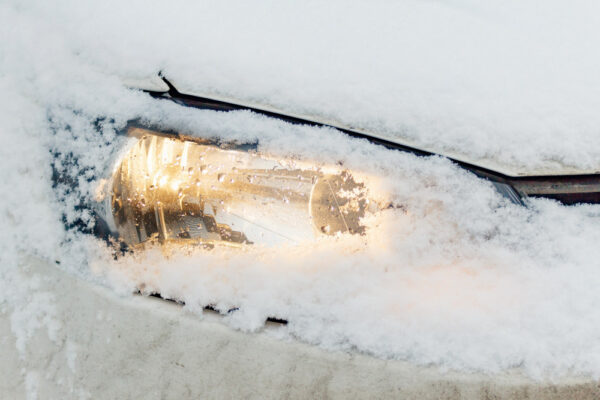 Eingeschneiter Autoscheinwerfer leuchtet durch die Schneedecke – typisches Bild an kalten Wintermorgen, wenn Autofahrer versucht sind, den Motor im Stand warmlaufen zu lassen