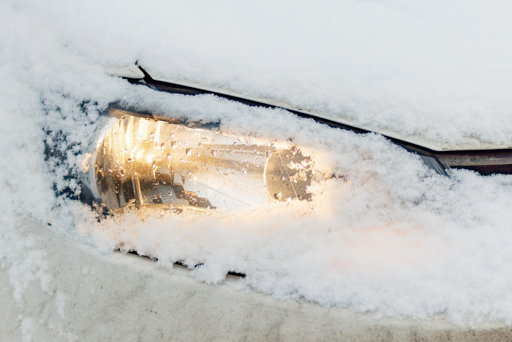Eingeschneiter Autoscheinwerfer leuchtet durch die Schneedecke – typisches Bild an kalten Wintermorgen, wenn Autofahrer versucht sind, den Motor im Stand warmlaufen zu lassen