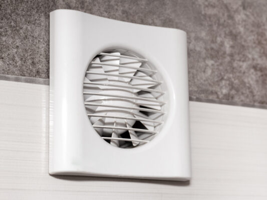 Ein weißer Wandlüfter mit rundem Schutzgitter sitzt auf einer hellen Wand unter einem grauen, rauen Wandbereich.