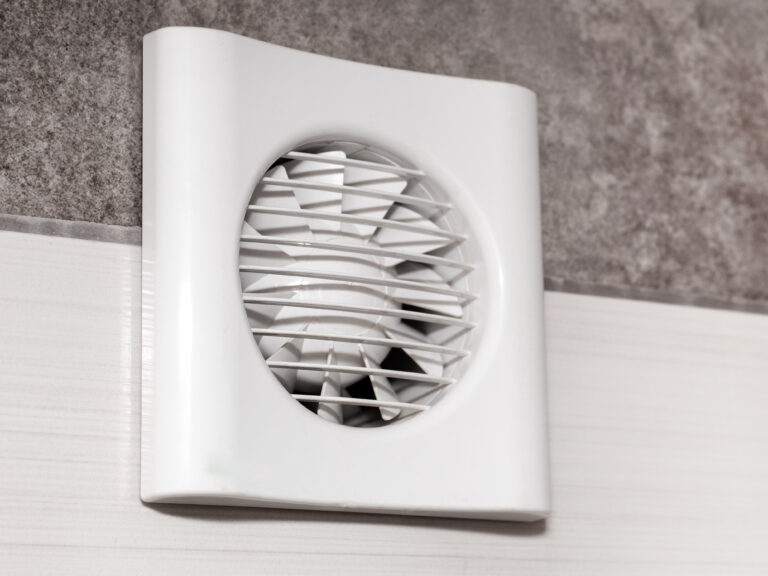 Ein weißer Wandlüfter mit rundem Schutzgitter sitzt auf einer hellen Wand unter einem grauen, rauen Wandbereich.