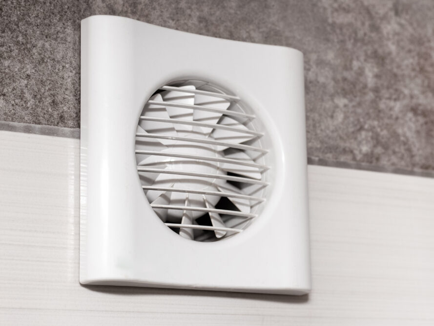 Ein weißer Wandlüfter mit rundem Schutzgitter sitzt auf einer hellen Wand unter einem grauen, rauen Wandbereich.