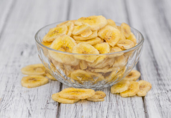 Gelbliche Bananenchips liegen in einer durchsichtigen Glasschale und verstreut auf einer hellen Holzfläche.