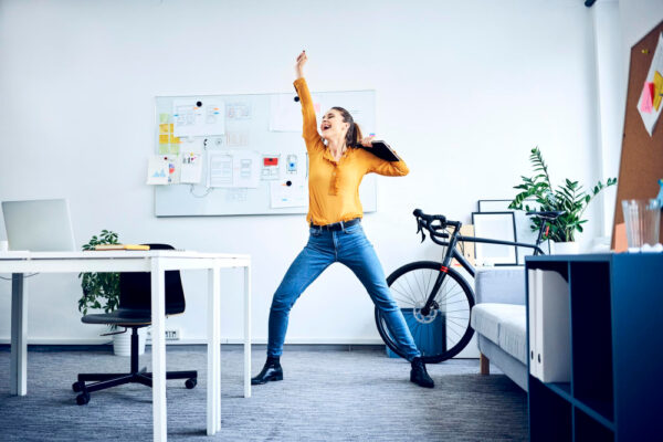Eine Frau in einem modernen Büro steht in einer dynamischen, jubelnden Pose, um die positive Energie und Freude durch mehr Bewegung am Arbeitsplatz zu symbolisieren.