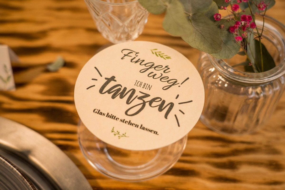 Bierdeckel selbst gestalten als runder Tischdeckel auf einem Glas, mit Spruch „Finger weg! Ich bin tanzen – Glas bitte stehen lassen“ auf einer festlich gedeckten Holztafel