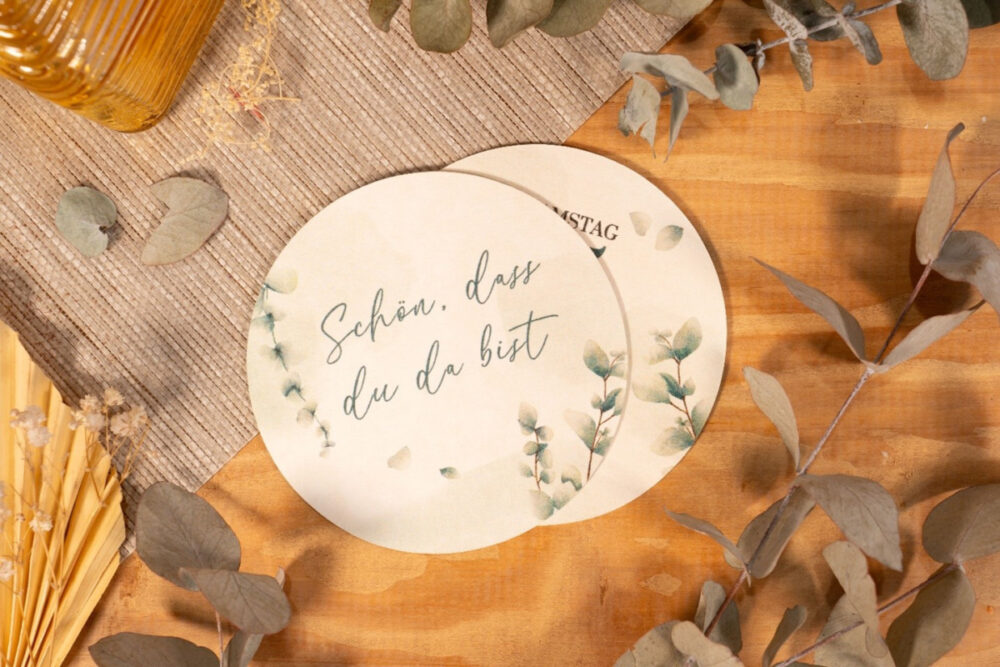 Bierdeckel selbst gestalten mit Aufdruck „Schön, dass du da bist“ im Eukalyptus-Design auf einem Holztisch mit Trockenblumen und Zweigen