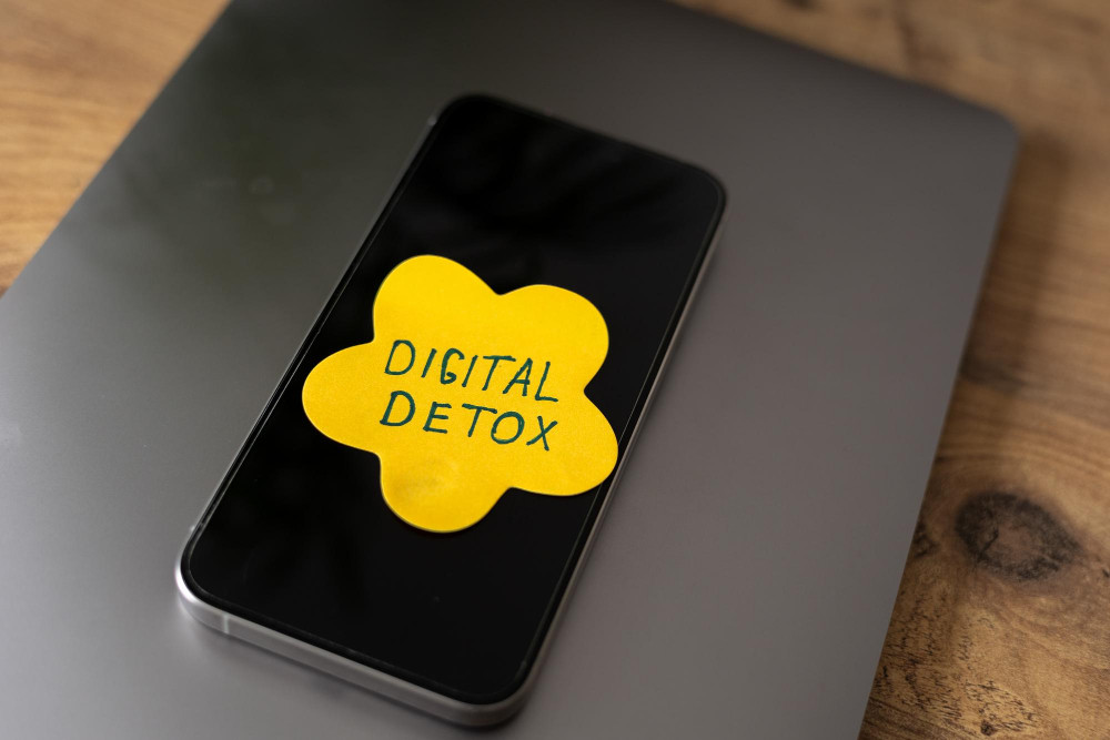 Ein schwarzes Smartphone liegt auf der geschlossenen, grauen Oberfläche eines Laptops. Auf dem dunklen Display des Handys klebt eine gelbe, blumenförmige Haftnotiz, auf der handschriftlich in dunkler Tinte die Worte "DIGITAL DETOX" stehen.