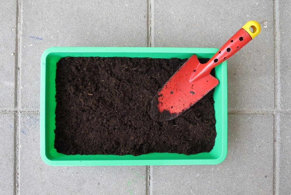 Grüner Pflanzkasten mit dunkler Blumenerde und roter Handschaufel auf Steinboden