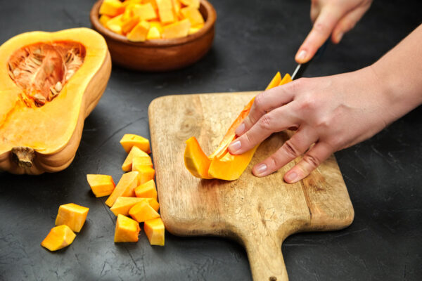 Hände schneiden mit einem Messer Butternut-Kürbis auf einem Holzbrett; daneben liegen eine Kürbishälfte und Würfel.
