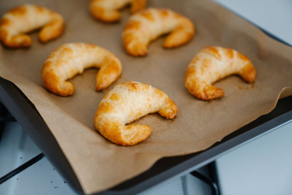 Mehrere goldbraune, halbmondförmige Croissants liegen auf braunem Backpapier auf einem dunklen Backblech.