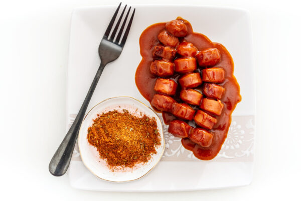 Auf einem weißen Teller liegen geschnittene Wurststücke in roter Soße, daneben stehen Currypulver und eine schwarze Gabel.