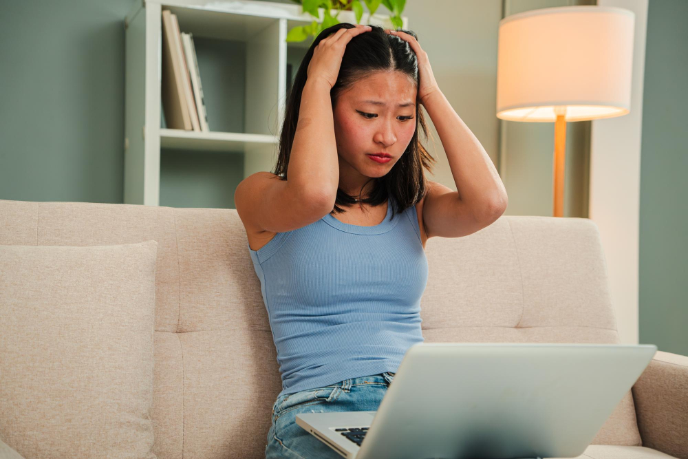 Junge Frau sitzt auf einem Sofa, hält sich den Kopf und sieht besorgt auf ihren Laptop, symbolisch für Stress und Sorgen, möglicherweise im Zusammenhang mit der Frage, ob der Arbeitgeber die Krankmeldung prüfen darf.