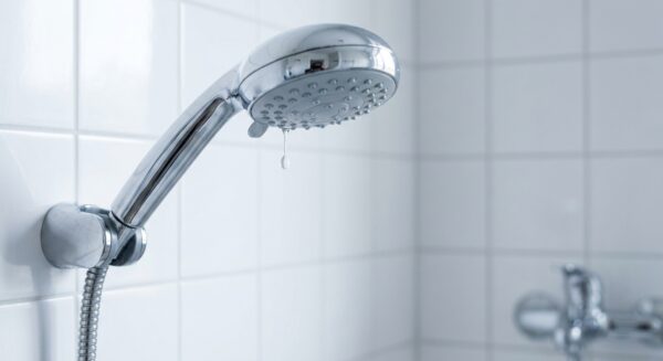 Ein moderner Duschkopf aus Chrom, montiert in einem weiß gefliesten Bad, aus dessen Düsen ein einzelner Wassertropfen langsam fällt.