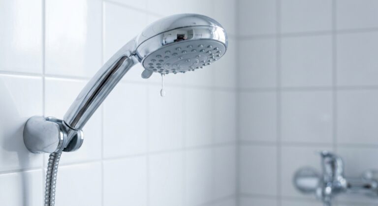 Ein moderner Duschkopf aus Chrom, montiert in einem weiß gefliesten Bad, aus dessen Düsen ein einzelner Wassertropfen langsam fällt.