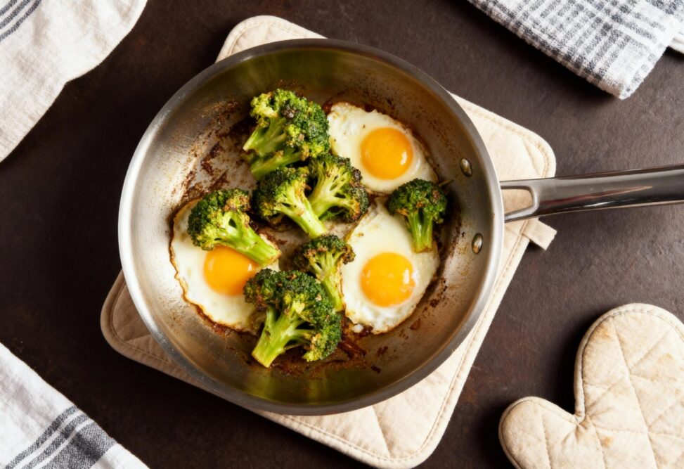 Edelstahlpfanne mit Spiegeleiern und geröstetem Brokkoli auf einem Topfhandschuh, daneben Küchentücher