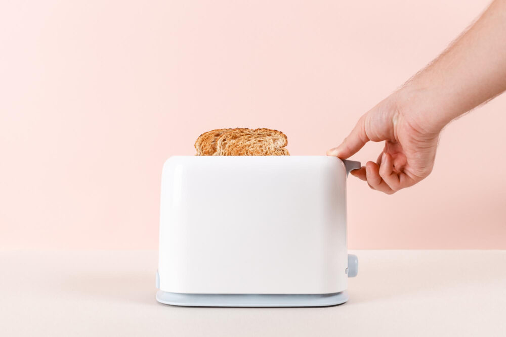 Hand drückt den Hebel eines weißen Toasters mit zwei Brotscheiben nach unten
