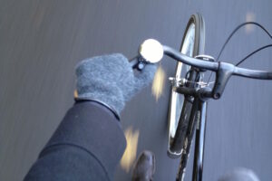 Eine Ich-Perspektive beim Fahrradfahren, die einen Arm im dunklen Mantel mit grauem Handschuh zeigt, der den Lenker eines schwarzen Fahrrads mit einer kleinen silbernen Klingel hält. Der Hintergrund ist unscharf, um Bewegung auf dem Asphalt anzudeuten.