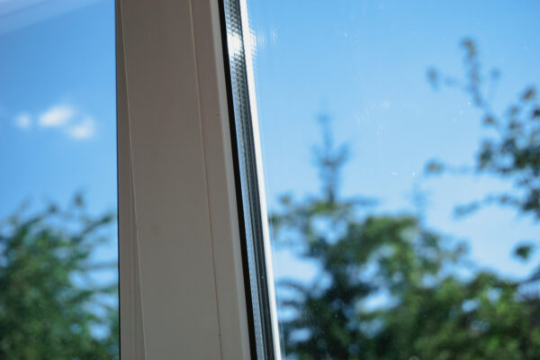 Nahaufnahme eines hellen Fensterrahmens mit Glasscheibe; dahinter sind blauer Himmel und unscharfe grüne Bäume zu sehen.