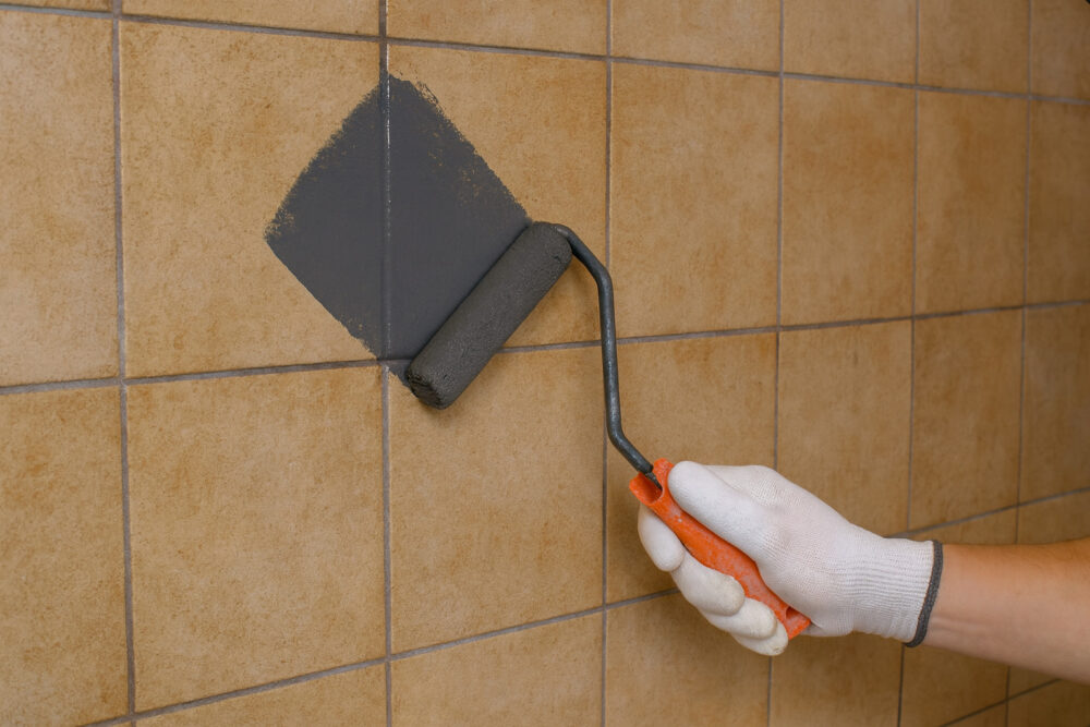 Hand mit Farbroller streicht alte beige-farbene Fliesen anthrazitfarben an