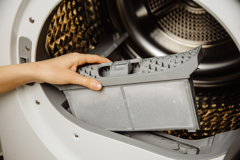 Eine Hand hält das graue Flusensieb vor der geöffneten Trommel eines Wäschetrockners.