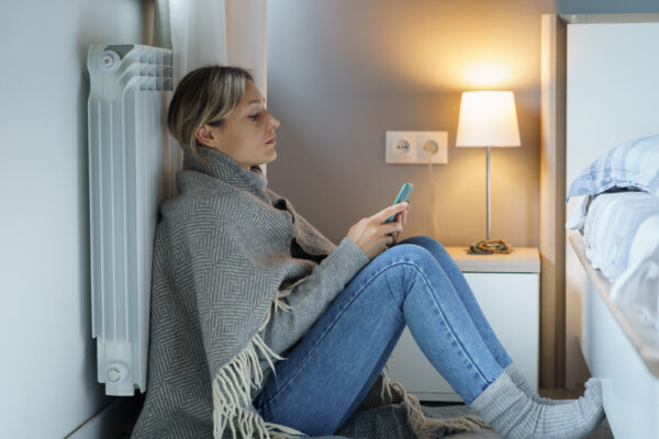 Eine Frau sitzt in Jeans und Decke neben einem weißen Heizkörper und schaut auf ihr Smartphone.