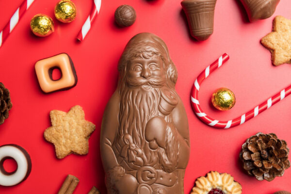 Frühe Weihnachtssüßigkeiten im Supermarkt: Ein großer Schokoladen-Weihnachtsmann liegt umgeben von weihnachtlichen Süßigkeiten wie Lebkuchen, Zuckerstangen, Plätzchen und Schokokugeln auf einem roten Hintergrund. Das Bild zeigt die Vielfalt an typischen Weihnachtssüßigkeiten, die bereits frühzeitig im Supermarkt erhältlich sind.