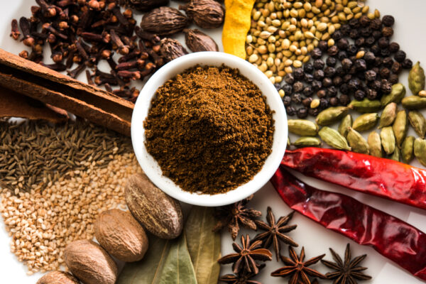Eine weiße Schale mit braunem Gewürzpulver steht zwischen Zimtstangen, Chilischoten, Kardamom und Pfefferkörnern.