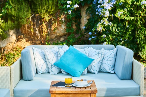 Helles Gartensofa mit blauen und gemusterten Kissen vor einer begrünten Steinmauer, davor ein Holztisch mit Geschirr.