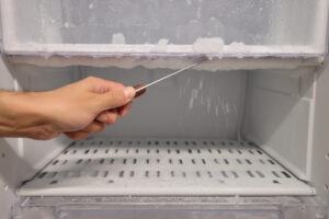Gefrierschrank richtig abtauen: Hand löst Eisschicht mit Schaber im Gefrierfach, Tropfwasser sichtbar