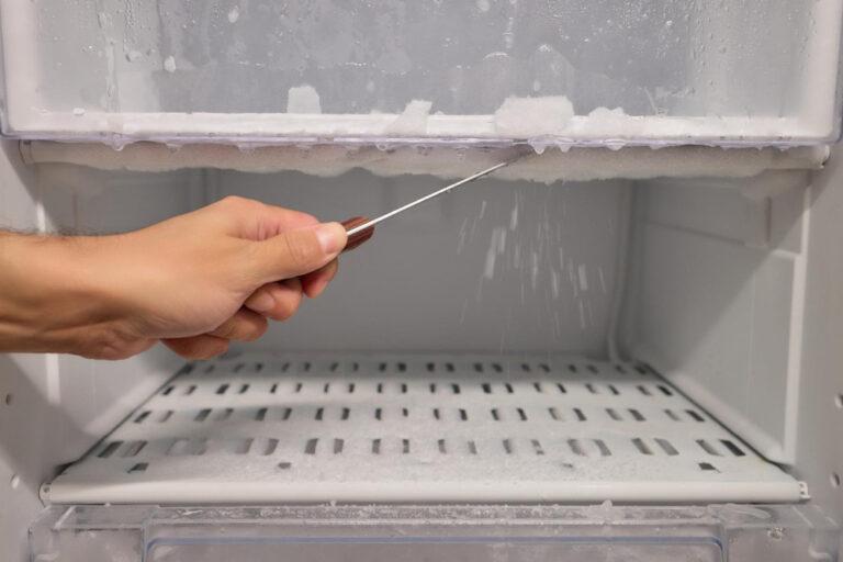 Gefrierschrank richtig abtauen: Hand löst Eisschicht mit Schaber im Gefrierfach, Tropfwasser sichtbar