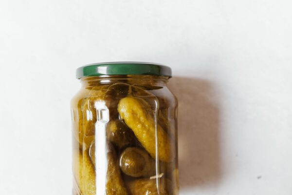 Ein verschlossenes Glas mit kleinen Gewürzgurken in gelblichem Sud steht vor hellem Hintergrund.