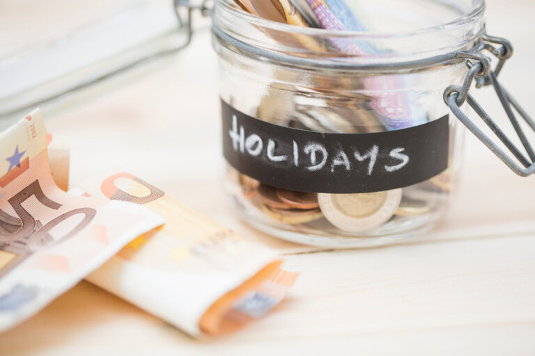 Glas mit Münzen und Geldscheinen, beschriftet mit „Holidays“, daneben Euro-Banknoten als Symbol für große Ausgaben vorbereiten