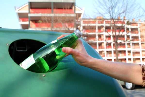 Eine Hand schiebt eine grüne Glasflasche durch die Öffnung eines grünen Altglascontainers.