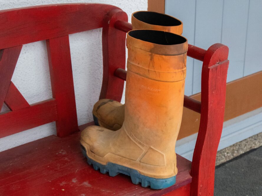 Ein Paar orangebraune Gummistiefel steht auf einer roten Holzbank neben einer hellen Tür.