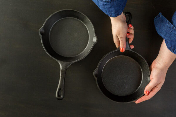 Zwei schwarze Gusseisenpfannen auf dunkler Arbeitsfläche, eine wird von Händen mit rotem Nagellack gehalten.