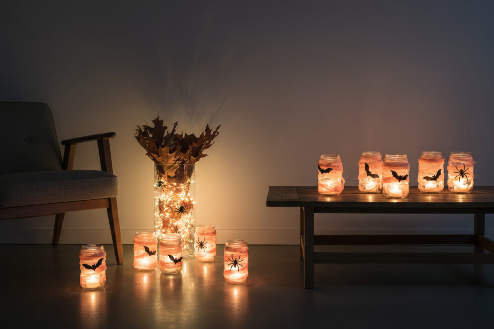 Stimmungsvolle Halloween-Beleuchtung in einem dunklen Raum. Mehrere Einmachgläser wurden zu Windlichtern umfunktioniert, die mit Fledermaus- und Spinnen-Silhouetten verziert sind und von innen warm leuchten.
