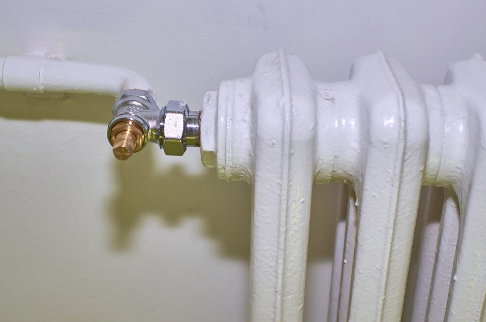 Nahaufnahme eines weißen Rippenheizkörpers mit metallischem Anschlussventil und weißem Rohr an einer hellen Wand.