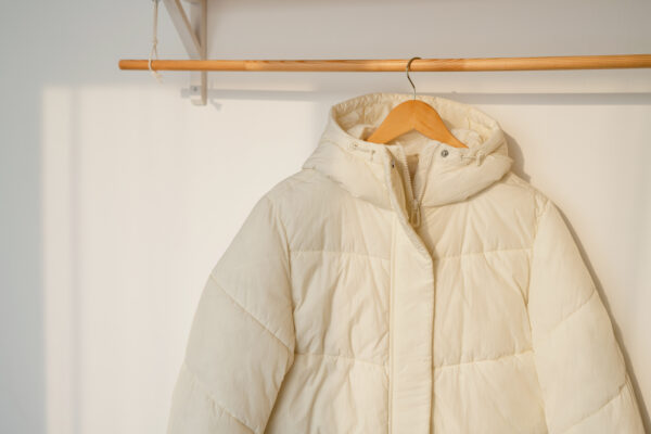 Eine helle, gesteppte Daunenjacke mit Kapuze hängt an einem Holzkleiderbügel an einer waagerechten Stange.