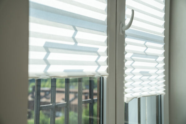 Zwei helle, gefaltete Plissees hängen vor einem Fenster; dahinter sind ein Geländer und Grün zu sehen.