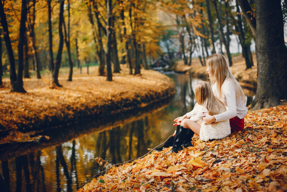 Mutter und Tochter sitzen am Flussufer im bunten Herbstlaub -stimmungsvolle Szene für einen Herbsturlaub in Deutschland.