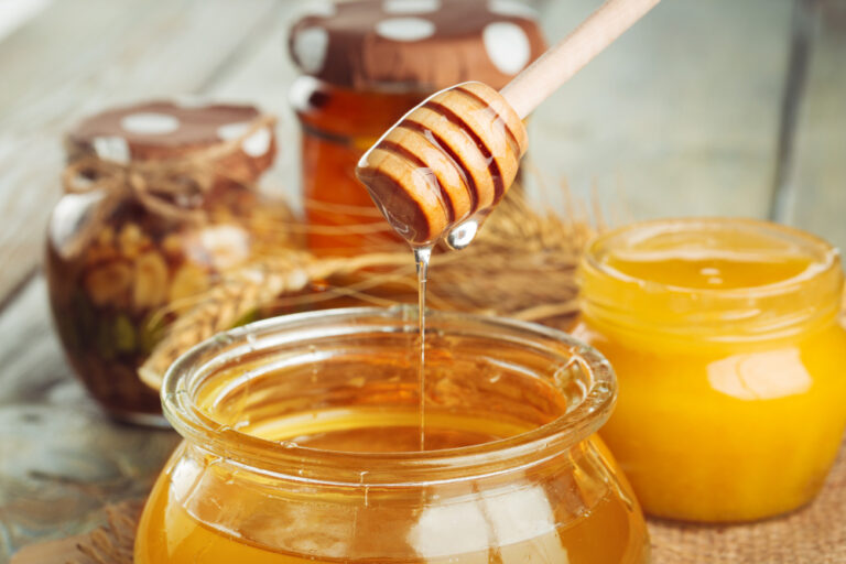 Holzlöffel lässt goldenen Honig aus einem offenen Glas fließen; weitere Honiggläser unscharf im Hintergrund - Thema: Honig wieder flüssig machen.