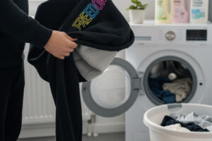 Eine Person dreht einen schwarzen Hoodie mit einem mehrfarbigen Aufdruck ("FREEDOM") auf links vor dem Waschen um, um den Aufdruck vor Abrieb zu schützen. Im Hintergrund ist eine offene Waschmaschine und ein Wäschekorb zu sehen.