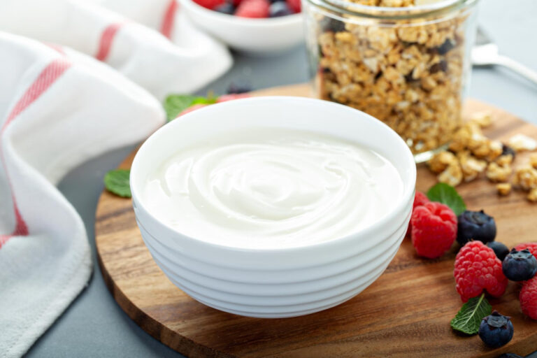 Schale mit cremigem selbst hergestelltem Joghurt auf einem Holzbrett mit Müsli, Himbeeren, Blaubeeren und Minze